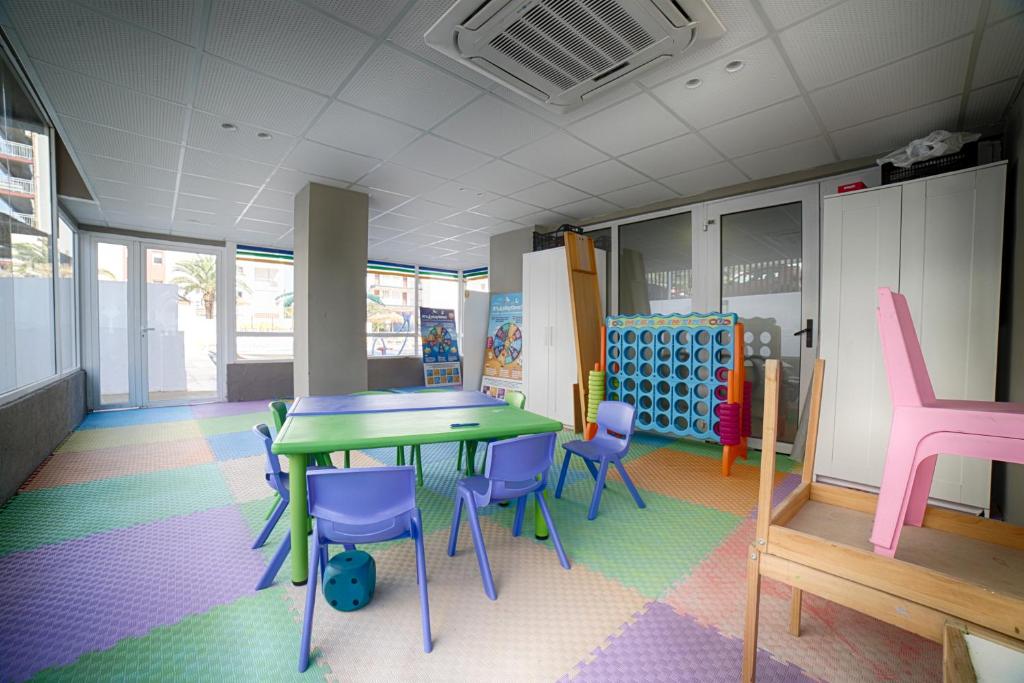 Bērnu rotaļu istaba ar krāsainām grīdām, zaļo galdu un zilu krēslu komplektu. Telpa ir gaiša un plaša, ar logiem, kas sniedz skatu uz ārpusi.