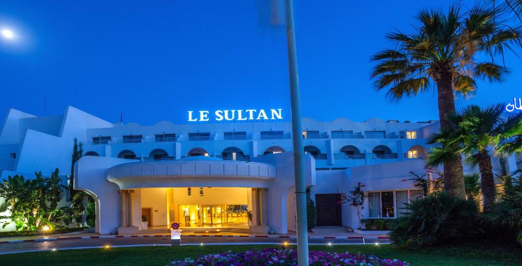 Viesnīcas "Le Sultan" ieeja naktī ar apgaismojumu un palmām apkārt. Skats uz eleganto arhitektūru un zilo debesu fonu.