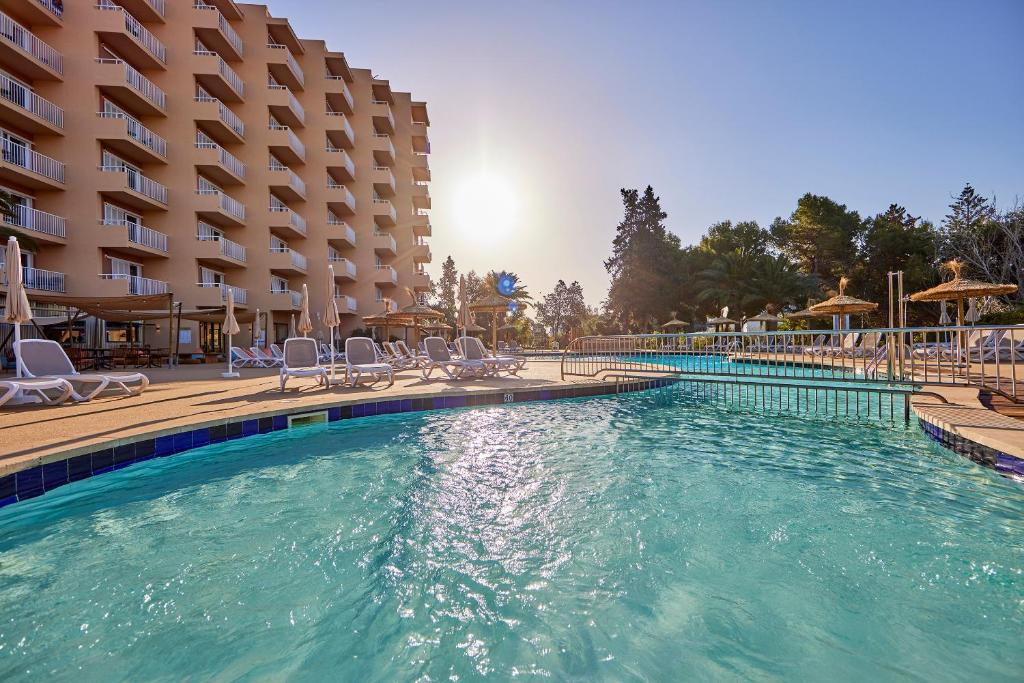 Sauļošanās baseins viesnīcā ar ērtām sauļošanās krēslām un koka jumtiem, kas izklāti ap baseinu, aizmugurē redzama viesnīcas ēka un saulriets.