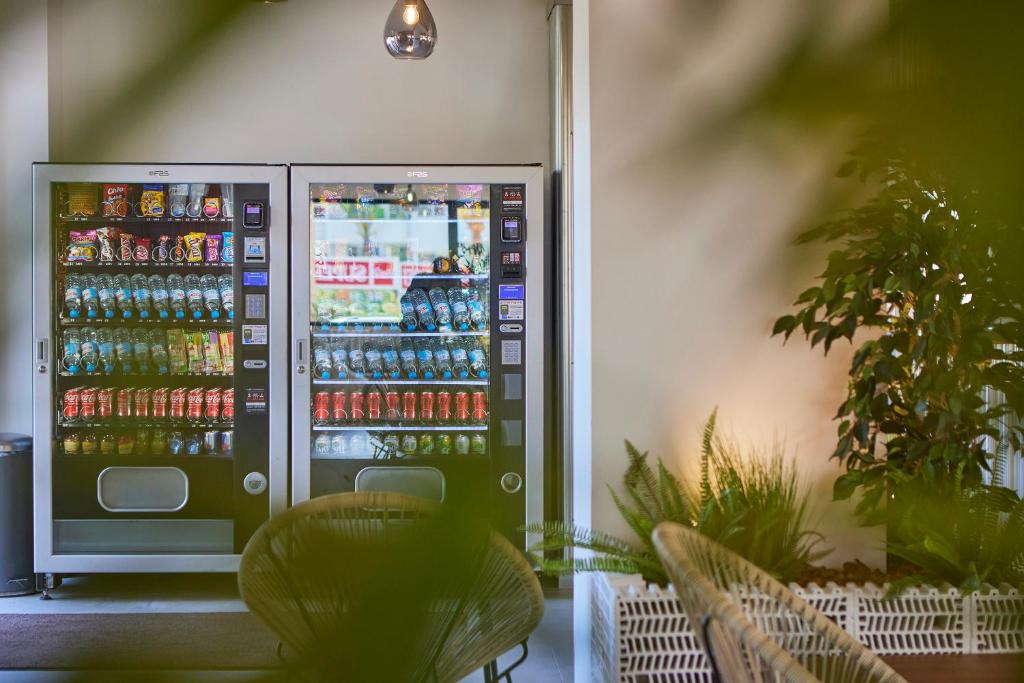 Automātu stends ar dzērieniem un uzkodām, atrodas mājīgā telpā ar zaļiem augiem un ērtām mēbelēm.
