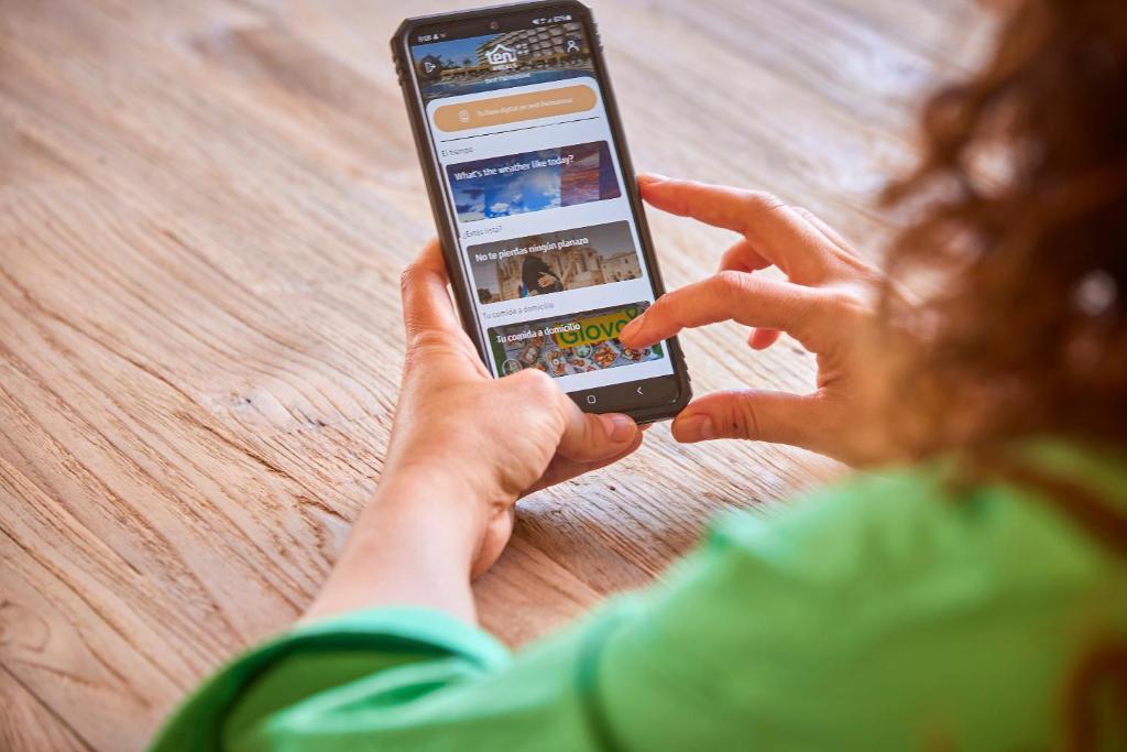 Personas roka tur mobilā tālruņa ekrānu, kurā redzama lietotne ar dažādiem interaktīviem elementiem. Fons ir koka galds.