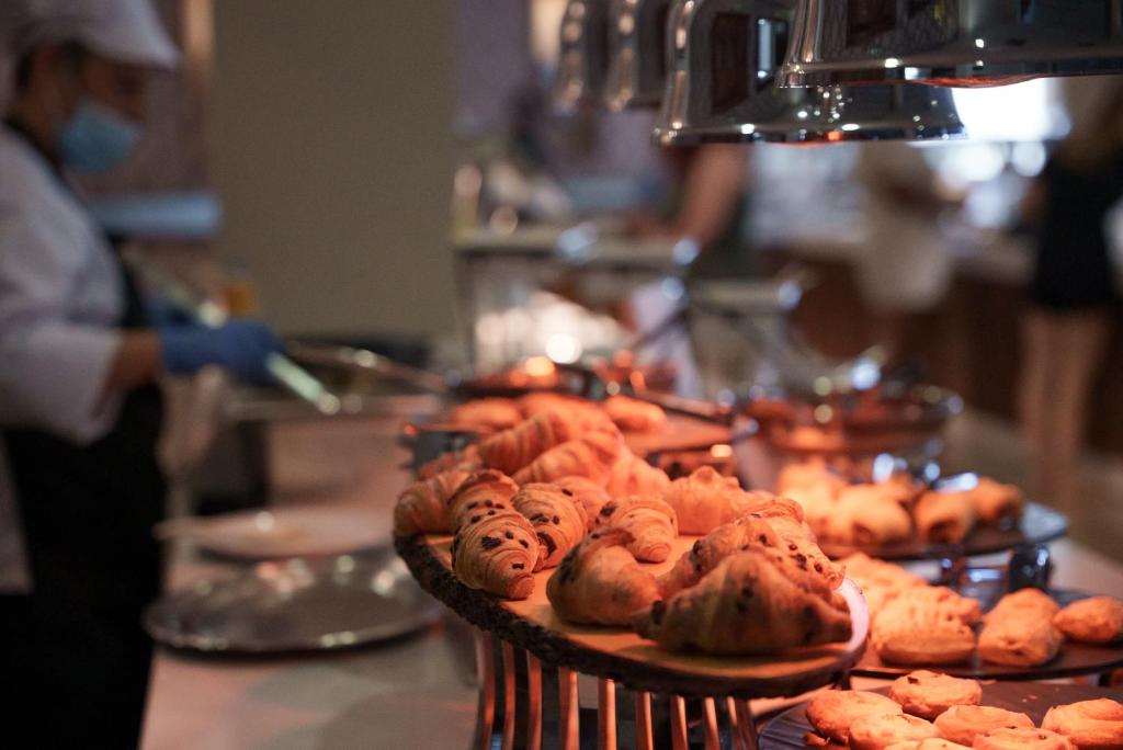Pusdienu bufete ar svaigiem kruasāniem un konditorejas izstrādājumiem, ko pasniedz šefpavārs. Apgaismojums rada siltu un viesmīlīgu atmosfēru.