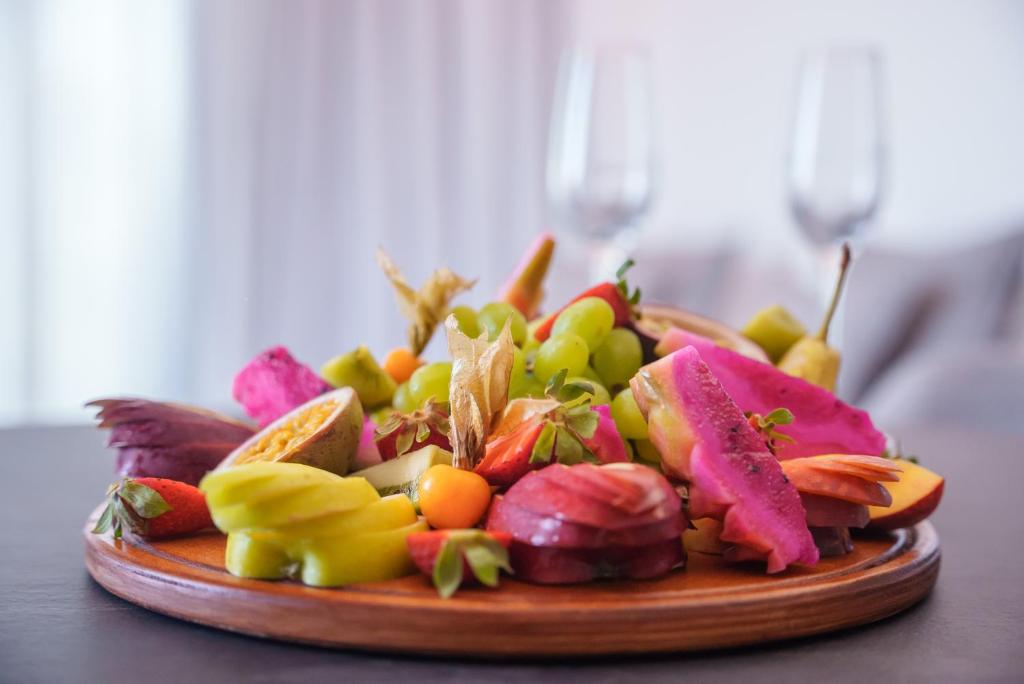 Krāsains augļu šķīvis ar dažādiem tropiskiem augļiem, piemēram, pitaju, vīnogām un zemenēm, pasniegts uz koka pamatnes. Fons ir mīksts un gaišs, radot patīkamu atmosfēru.