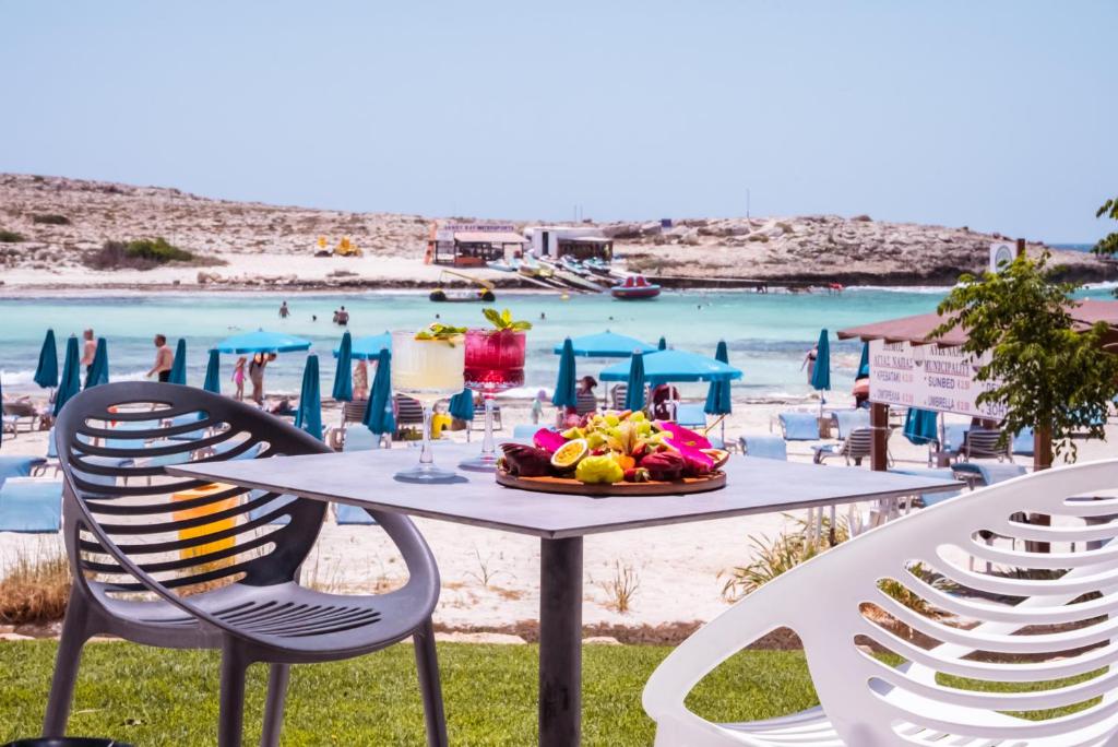 Pludmales restorāna skats ar augļu šķīvi uz galda un zilu ūdeni fonā, kur cilvēki atpūšas zem saules. Ideāla vieta atpūtai un relaksācijai.