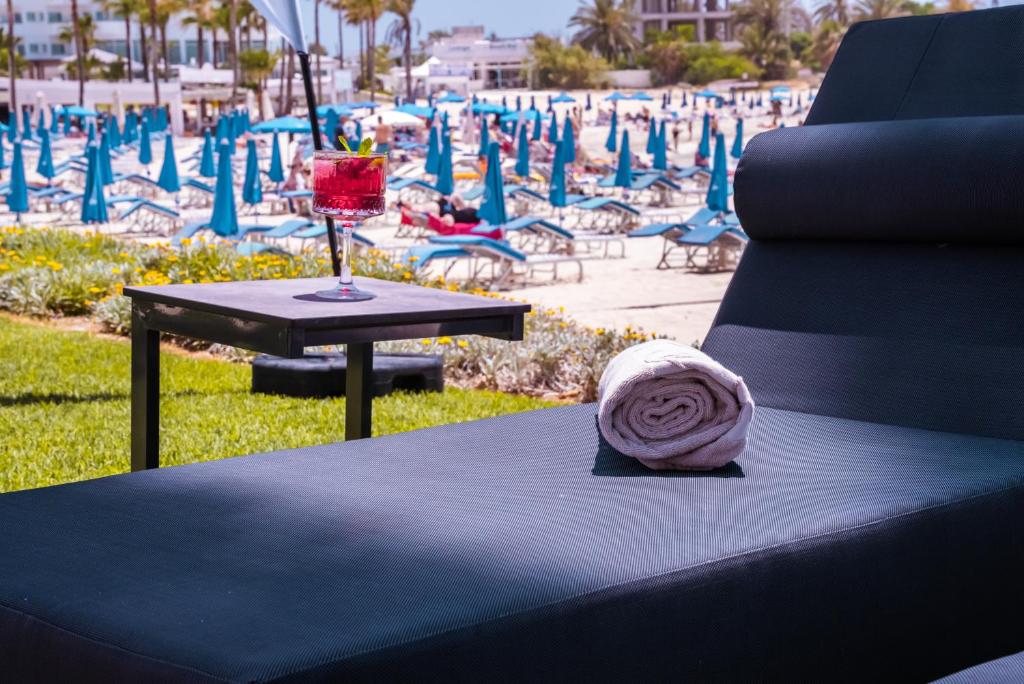 Saulainā pludmales atpūtas zona ar šikiem sauļošanās krēsliem, galdiņu un dzērienu, kas sagatavo viesiem relaksējošu pieredzi. Apkārt redzamas zila krāsā nokrāsotas sauļošanās parasoles un pludmales atmosfēra.
