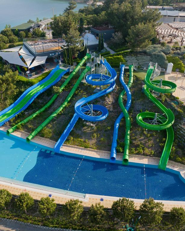 Ūdens atrakciju parks ar dažādiem slaidiem, kas izvietoti kalnā blakus peldbaseinam. Skats no augšas uz zaļajiem un ziliem slaidiem, kas piedāvā jautras izklaides iespējas.