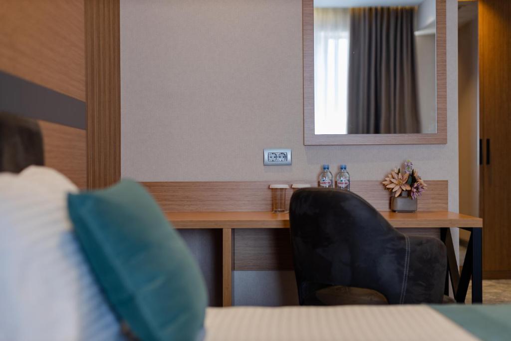 Mūsdienīgs viesnīcas numurs ar koka mēbeļu elementiem, galdu un spoguli. Uz galda redzamas ūdens pudeles un ziedi, kas piešķir telpai mājīgumu.