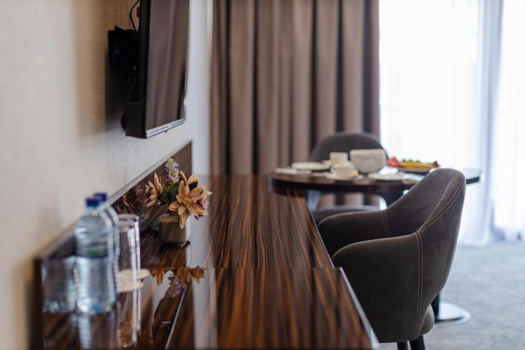 Mūsdienīgs viesnīcas numurs ar elegantu koka galdu, televizoru pie sienas un dekoratīvām puķēm. Galdā ir pasniegti ēdieni, un blakus redzami ērti krēsli.