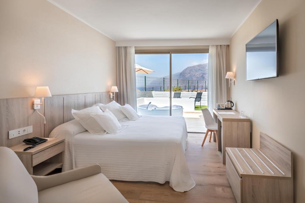 Mūsdienīga viesnīcas numura iekārtojums ar gaisīgu interjeru, ērtu gultu un skatu uz terasi. Logi ļauj ieplūst dabiskai gaismai, radot patīkamu atmosfēru.