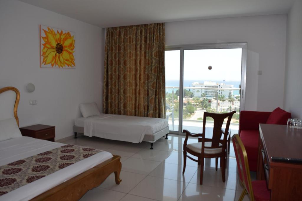 Mūsdienīgs viesnīcas numurs ar divām gultām, koka mēbelēm un gaisīgu interjeru, kas piedāvā skatu uz jūru. Logi ar aizkariem un saulespuķu glezna uz sienas piešķir telpai siltumu.