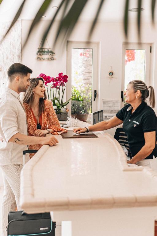Reģistratūras darbinieks sveicina viesus, kuri priecīgi sarunājas pie viesnīcas reģistratūras. Interjerā redzami ziedi un gaiša atmosfēra.