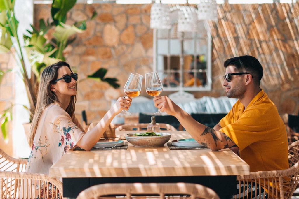 Pāris, kas sēž restorānā, paceļ glāzes, svinot mirkli. Gaisīgā un mājīgā atmosfērā ir redzami zaļumi un elegantas mēbeles.