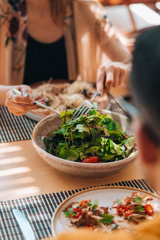 Veselīga salātu ēšana pie galda, ar dāmām, kas izmanto dakšiņas, lai pasniegtu zaļumus. Gaisma plūst cauri logam, radot patīkamu atmosfēru ēdienreizei.