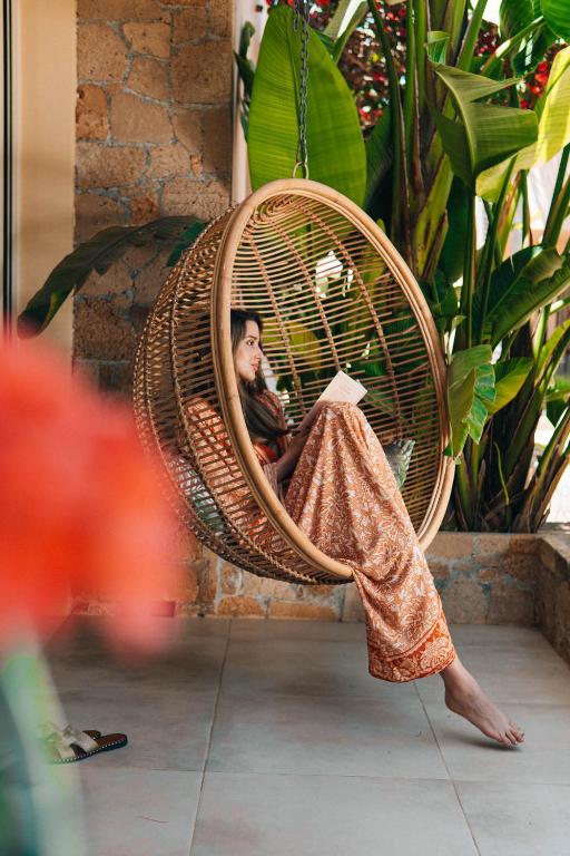 Sieviete, kas sēž šūpuļtīklā, lasa grāmatu, apkārt aug zaļas palmas un ziedi, radot mierīgu atmosfēru.