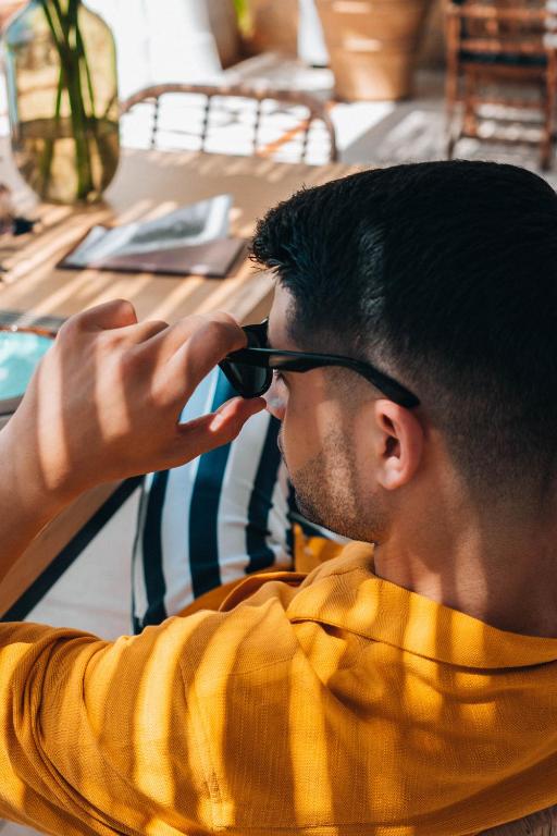Jauns vīrietis, tērpts dzeltenā krekliņā, pielāgo saulesbrilles, sēžot modernā interjerā. Gaisma veido interesantas ēnas uz viņa sejas.