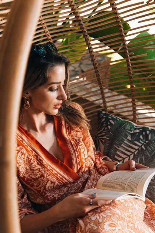 Sieviete, tērpta sarkano toņu kleitā, lasa grāmatu ērtā sēžamvietā ar augu fonu. Daba un relaksācija apvienojas harmoniskā atmosfērā.