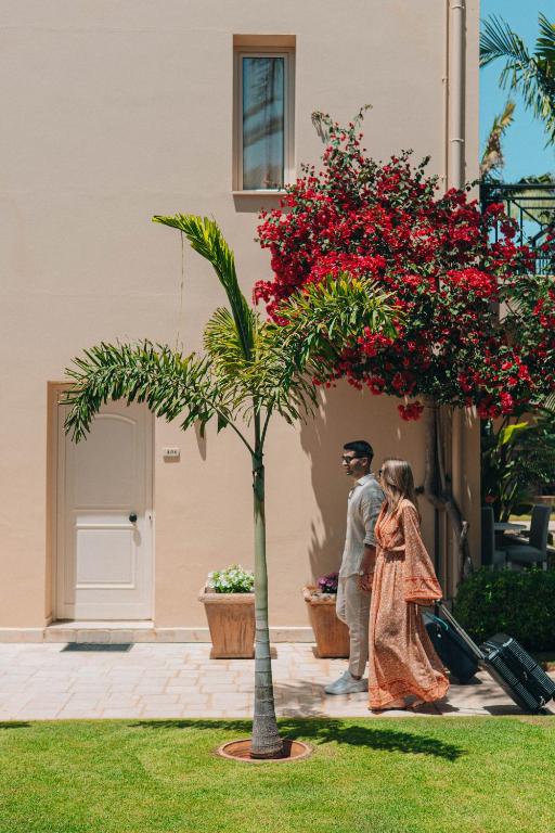 Pāris pastaigā pa dārzu, kamēr viņi velk ceļojuma somas. Fons ir mājvieta ar ziediem un palmām.