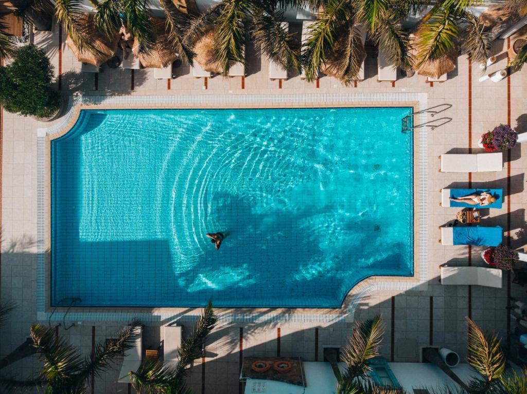 Baseins ar skaidru ūdeni un palmām ap to, kurā cilvēks peld. Apkārt baseinam redzamas saulessargu un gultu rindas, radot atpūtas atmosfēru.