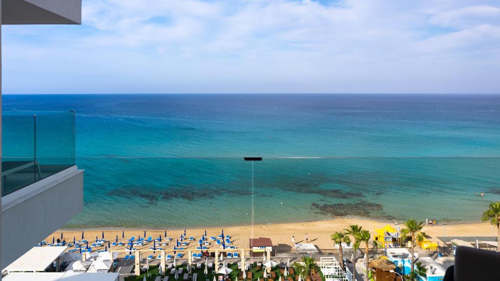 Skats uz jūru no viesnīcas balkona, kur redzama smilšu pludmale ar saulessargiem un zilo ūdeni, kas saplūst ar debesīm. Ideāla vieta atpūtai un relaksācijai.