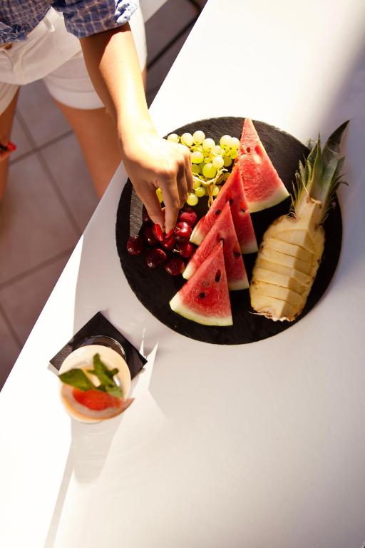 Svaigu augļu šķīvis ar arbūziem, vīnogām, ķiršiem un ananāsu, ko kāds grasās pamēģināt. Blakus ir kokteilis ar zemeņu un piparmētru dekoru.