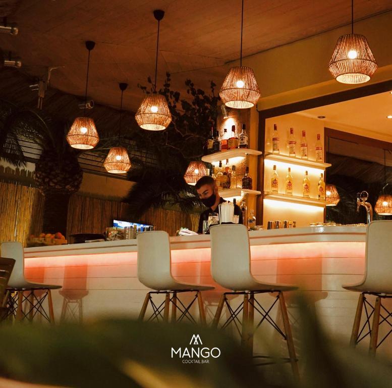 Mango kokteiļu bārs ar modernu interjeru, kur bārmenis sagatavo dzērienus pie eleganta apgaismotā bāra. Siltas gaismas un tropiskas augu detaļas rada patīkamu atmosfēru.