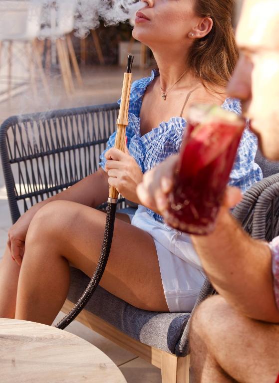 Sieviete, kas sēž uz terases, turot rokās ūdenspipes cauruli un izpūšot dūmus, blakus viņai ir vīrietis, kas tur sarkanu dzērienu. Fonā ir moderna kafejnīcas vide.