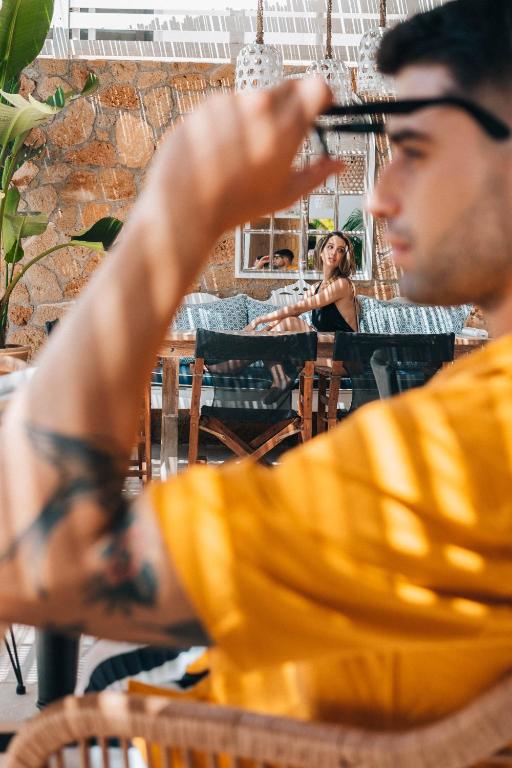 Jaunā stilīgā kafejnīcā redzams jauns vīrietis, kurš skatās uz sievieti, kas sēž fonā. Vīrieša tērps ir spilgti dzeltenā krāsā, bet sieviete ir apģērbta melnā. Gaisma rada patīkamu atmosfēru, un telpa ir iekārtota ar koka mēbelēm un zaļām augu dekorācijām.