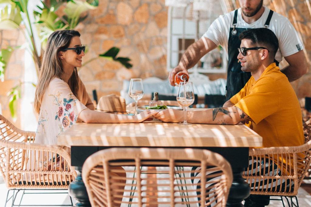 Romantiska maltīte diviem modernā restorānā, kurā viesmīlis pasniedz dzērienu. Vīrietis un sieviete ir ģērbušies stilīgi un valkā saulesbrilles, sēžot pie koka galda.