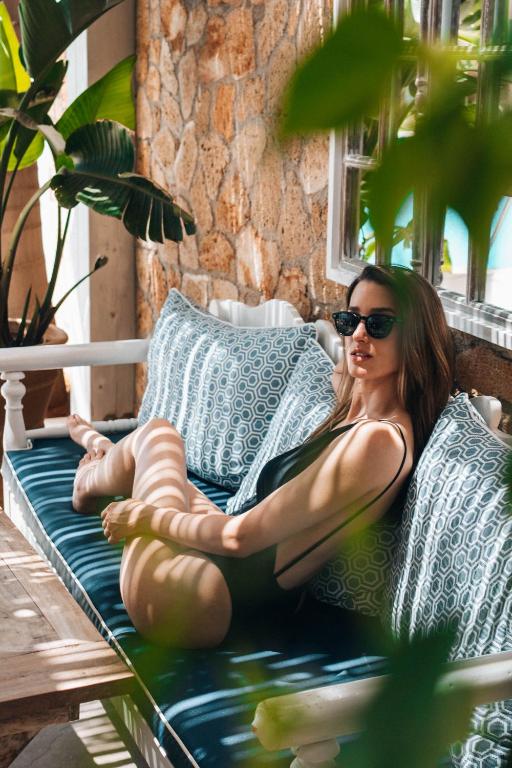 Sieviete brīvdienās atpūšas uz dīvāna, valkājot peldkostīmu un saulesbrilles, ar zaļiem augiem un akmens sienu fona.