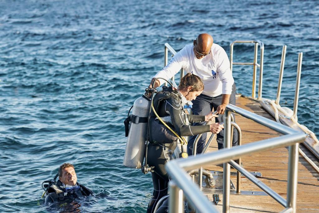 Duviniekam sagatavojoties niršanai, viens cilvēks palīdz otram uzvilkt niršanas aprīkojumu uz piestātnes, kamēr trešais atrodas ūdenī.