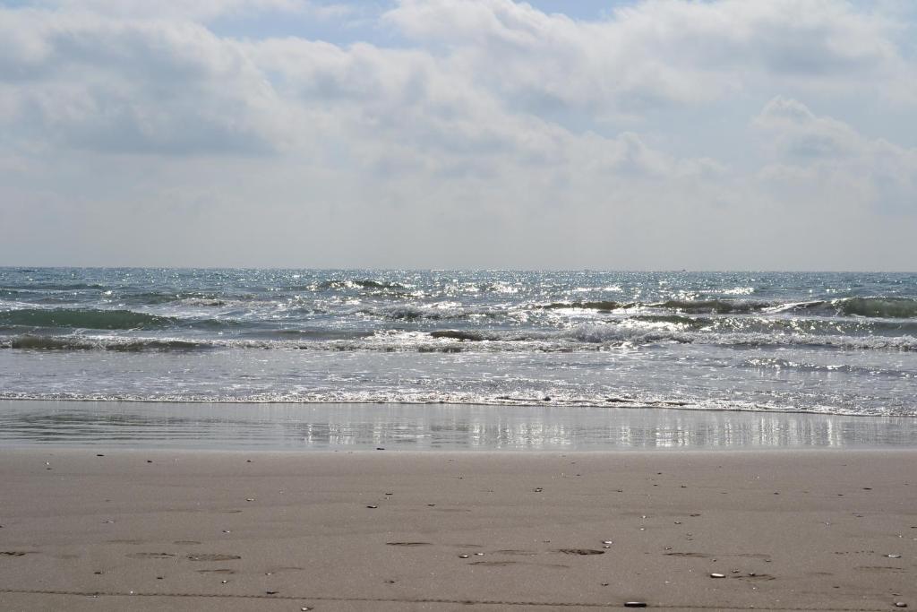 Jūras ainava ar viļņiem un saules staru atspulgus uz ūdens virsmas, kas rada mierīgu atmosfēru. Pludmale ir klāta ar smiltīm un nelieliem akmentiņiem.