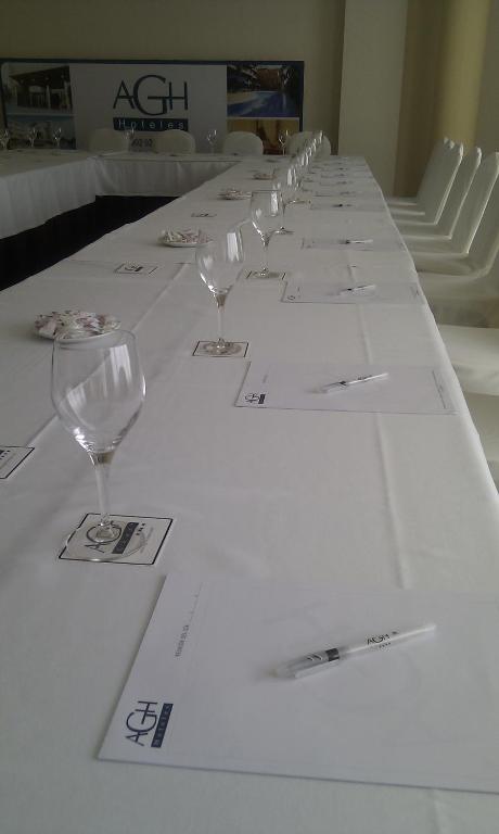 Konferenču telpa ar garu galdu, kas klāts ar baltu galdautu, uz kura ir glāzes, piezīmju lapas un pildspalvas. Fons satur AGH viesnīcu logo un attēlus.