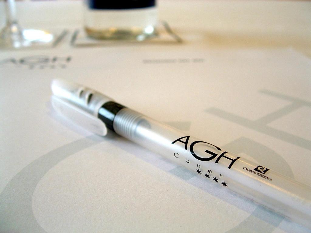 Balta pildspalva ar melnu uzrakstu "AGH Canet" uz papīra, kas atrodas uz galda. Fons ir izplūdis, radot profesionālu atmosfēru.