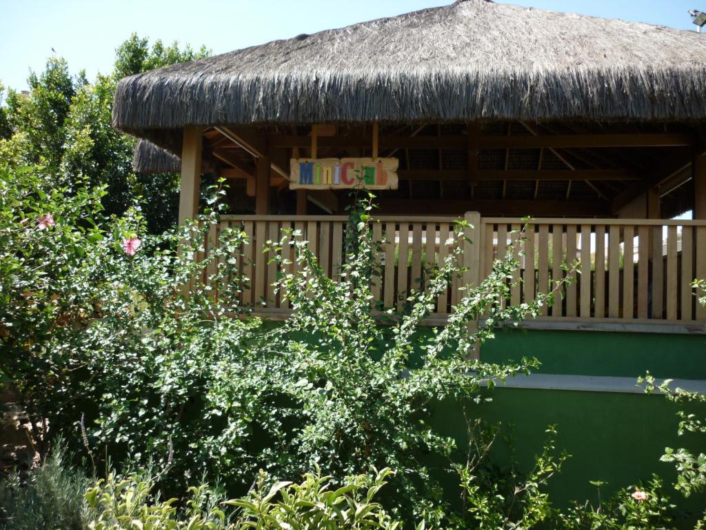 Bērnu klubs ar salmu jumtu un krāsainu zīmi, ieskauts zaļiem augiem un ziediem, rada draudzīgu un aicinošu atmosfēru.