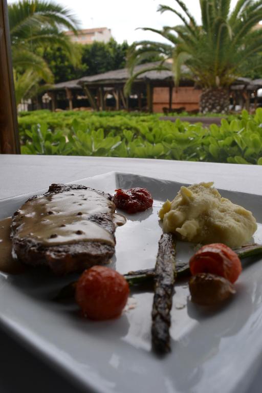 Grilēta gaļa ar mērci, apkalpota ar kartupeļu biezeni, grauzdētām tomātiem un sparģeļiem, ar zaļām palmām fona.