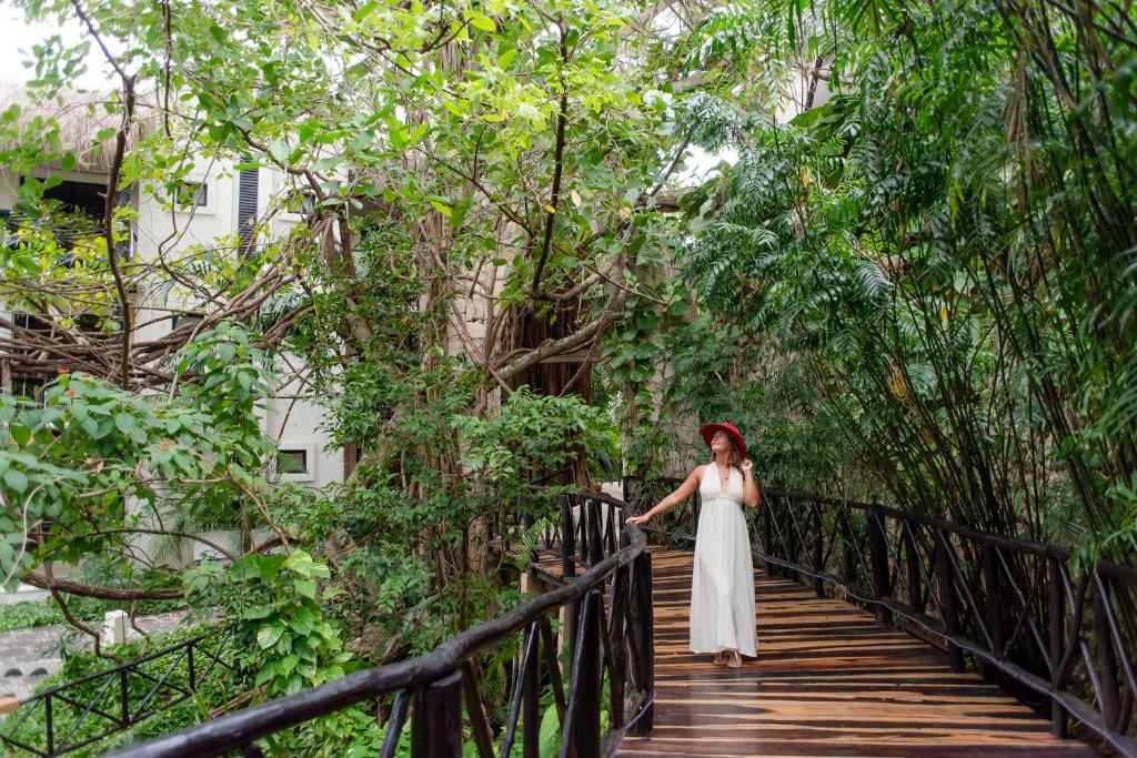 Sieviete baltā kleitā un sarkānā cepurē pastaigā pa koka tiltu, ko ieskauj zaļa veģetācija un koku zari. Daba un mierīga atmosfēra veido harmonisku ainu.