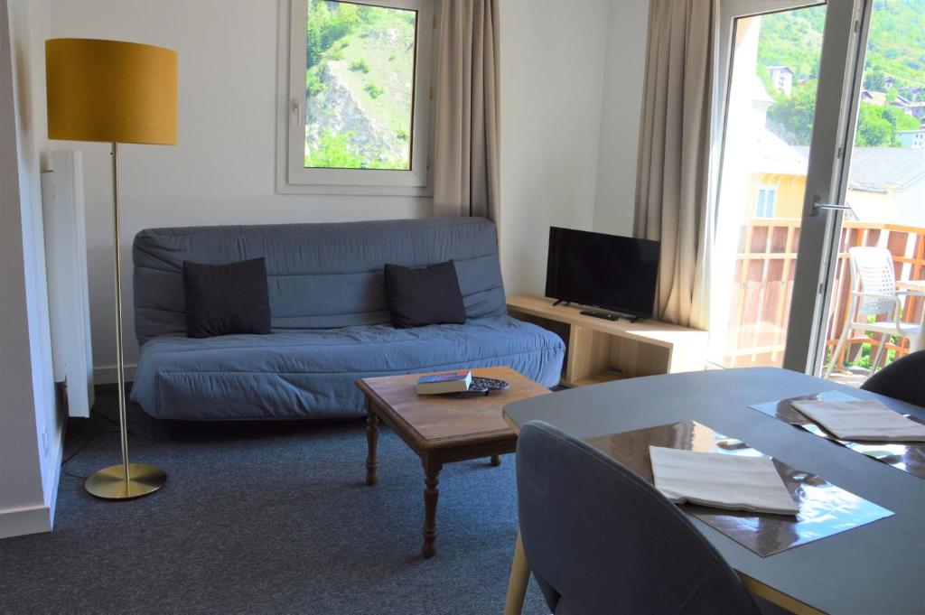 Mūsdienīgs dzīvojamās istabas interjers ar dīvānu, zelta krāsas grīdas lampu un televizoru, kas izskatās uz balkonu ar zaļo skatu.