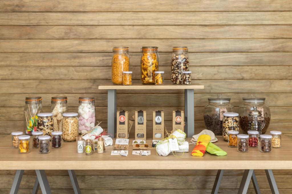 Dažādu uzkodu un garšvielu burkas uz koka galda, radot krāsainu un pievilcīgu izskatu. Burkas satur riekstus, saldumus un citas uzkodas, papildinātas ar dekoratīviem etiķetēm.