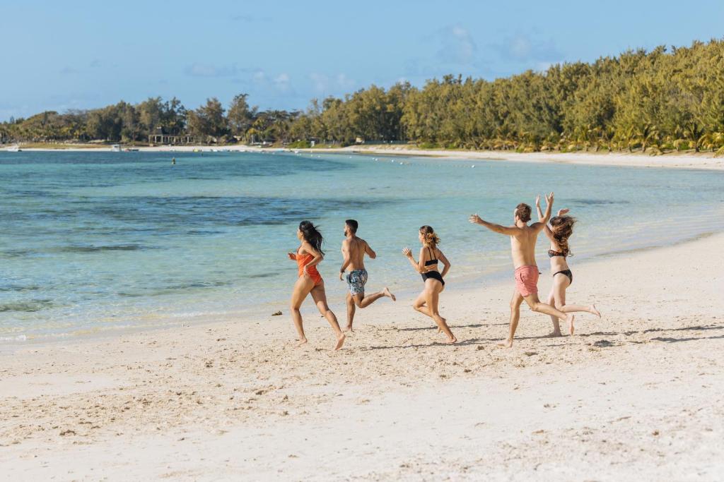 Grupa cilvēku izbauda pludmales aktivitātes, skrienot gar smilšaino krastu. Fons ir skaista jūras ainava ar palmām un saulainu debesi.