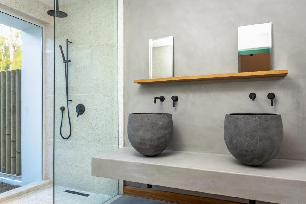 Mūsdienīgs vannas istabas interjers ar divām apaļām betona izlietnēm, koka plauktu un melnu santehniku. Gaismas un tekstūras saskaņa rada elegantu un minimālistisku atmosfēru.