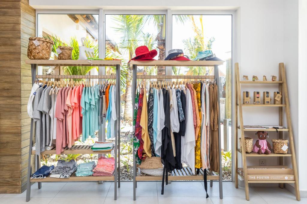 Veikala interjers ar divām drēbju plauktiem, kas pildīti ar krāsainām t-krekliem un biksēm, kā arī aksesuāriem, piemēram, cepurēm un groziem. Logi sniedz skatu uz zaļu dārzu ar palmu kokiem.