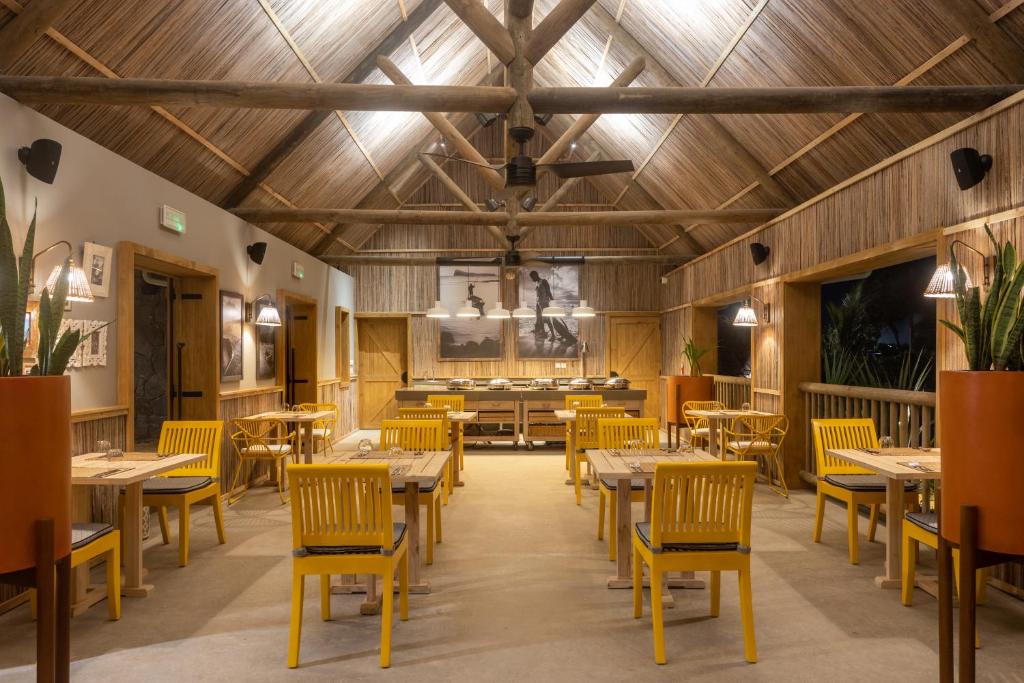 Mūsdienīga restorāna telpa ar koka elementiem, dzeltenām krēsliem un siltu apgaismojumu, radot viesmīlīgu atmosfēru. Interjerā redzami mākslas darbi un augi, kas papildina dizainu.