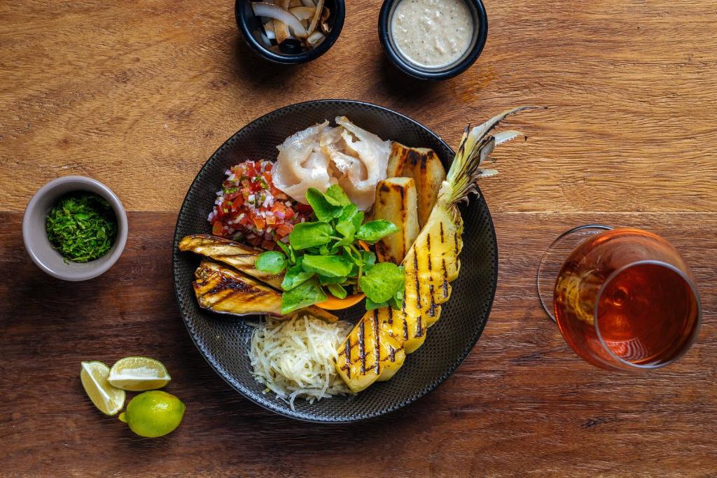 Krāsains ēdiena šķīvis ar grilētu ananāsu, dārzeņiem un mērci, blakus citrona šķēlītes un dzēriens. Ideāls piemērs veselīgai un garšīgai maltītei.