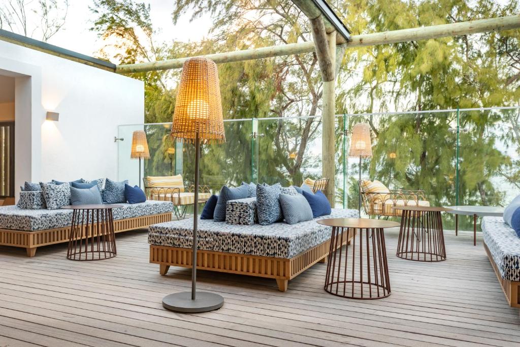 Mūsdienīgs terases dizains ar ērtām mēbelēm, kas ietver dīvānus ar spilveniem un stilīgas lampas. Telpa ir apgaismota ar dabisku gaismu un ir ieskauta zaļiem kokiem.