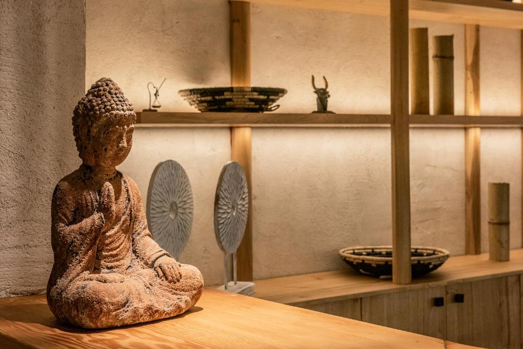 Buddhas statuja, kas sēž uz koka galda, priekšplānā, ar mūsdienīgu fona interjeru un dekoratīviem priekšmetiem plauktos.
