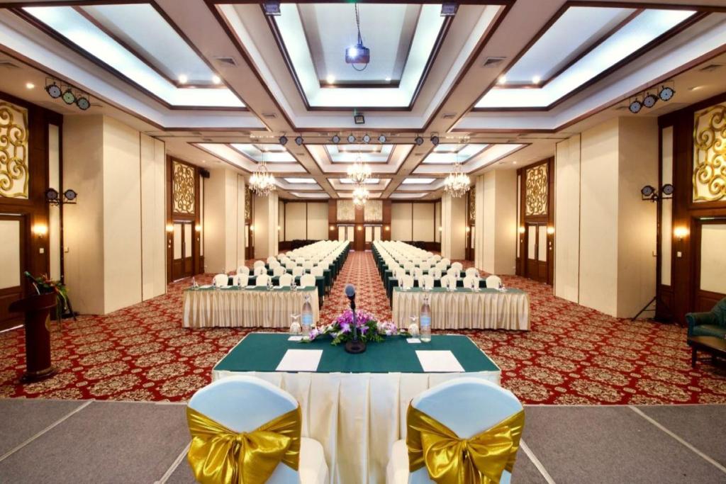 Konferenču zāle ar rindu galdiem un krēsliem, dekorēta ar ziediem un apgaismojumu. Eleganta vide piemērota pasākumiem un sanāksmēm.