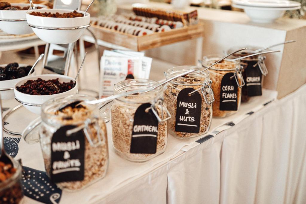 Brokastu bufete ar dažādiem graudaugiem un riekstiem stikla burkās, kas marķētas ar melniem uzrakstiem. Galds ir rotāts ar baltu galdautu un citiem ēdieniem.