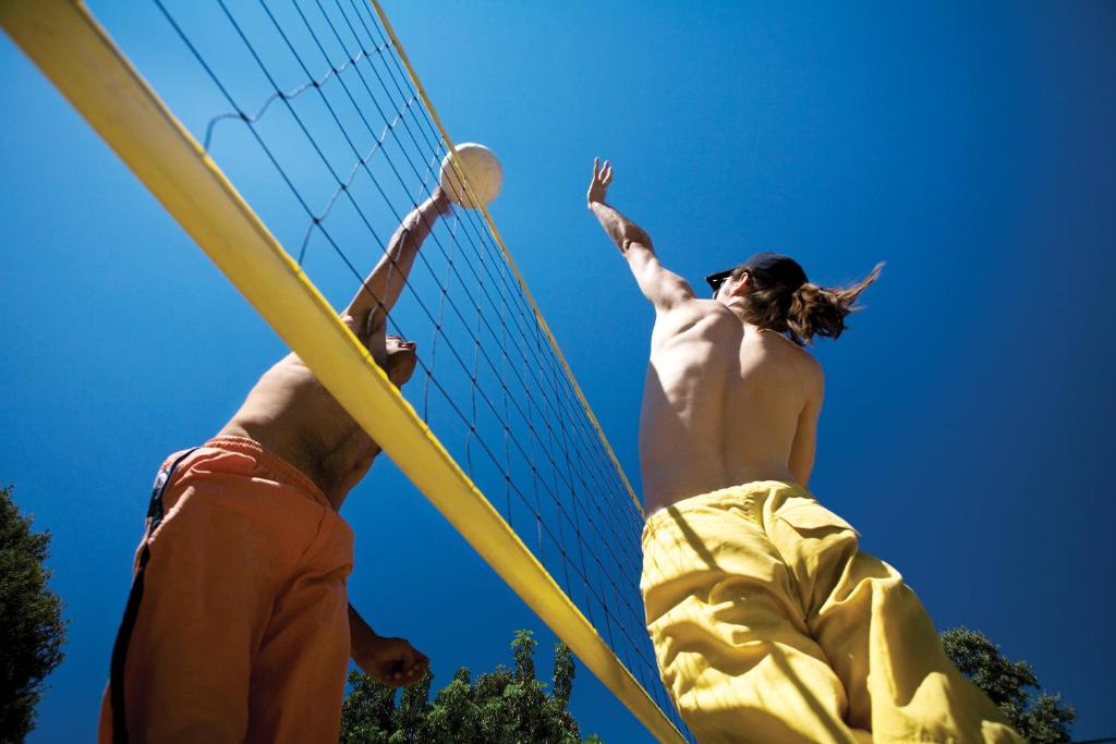 Divi cilvēki spēlē pludmales volejbolu zem skaidra zila debesīm, centīgi uzsitot bumbu pāri tīklam. Spēles dinamika un enerģija ir acīmredzama.