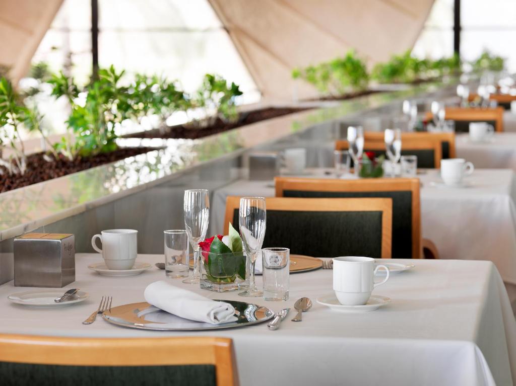 Eleganta restorāna galda uzstādīšana ar ziediem centrā un stilīgiem traukiem. Gaisīga atmosfēra ar zaļumiem un modernu dizainu.