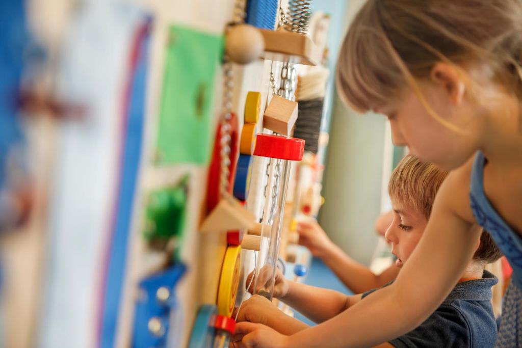 Bērni spēlējas ar interaktīvo mācību paneli, kurā ir daudzkrāsaini elementi, kas attīsta viņu motoriku un radošumu.